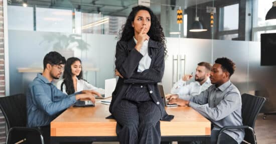 Abbildung: Eine Frau im Business-Outfit sitzt nachdenklich auf einem Tisch in einem Büro, an dem Kolleg*innen mit Notebooks sitzen.