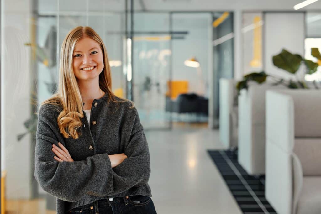 Alina steht mit verschränkten Armen lächelnd angelehnt an eine Glaswand im 247GRAD Office.