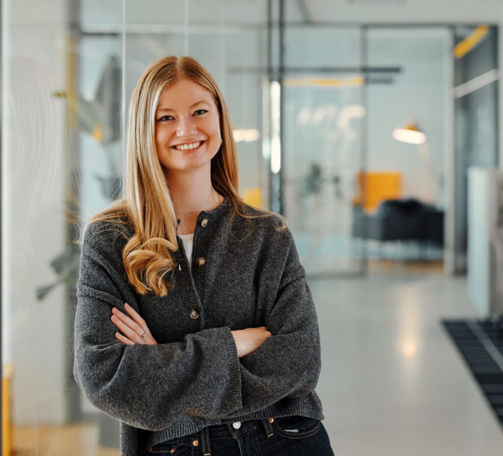 Alina steht mit verschränkten Armen lächelnd angelehnt an eine Glaswand im 247GRAD Office.