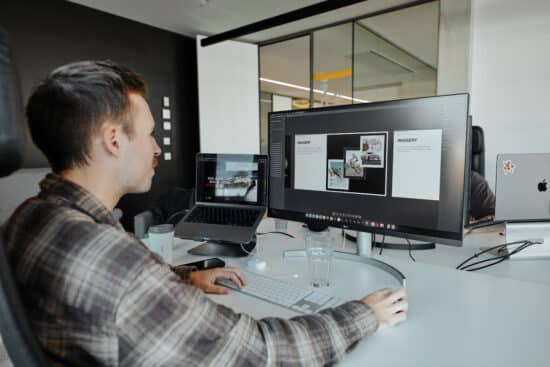 Noah ist an einem Schreibtisch und hat dort seinen Laptop und einen weiteren Bildschirm, auf dem er Designelemente für ein Kundenprojekt entwirft. Er ist von hinten zu sehen und schaut auf den Bildschirm, die Hand an der Maus.