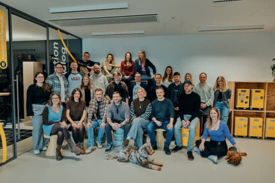 Das Team von 247GRAD sitzt und steht in mehreren Reihen im Office. Insgesamt sind 25 Personen zu sehen und zwei Bürohunde. Alle haben unterschiedliche Kleidung an und lächeln in die Kamera. Im Hintergrund sieht man einen Besprechungsraum und rollbare Flightcases in einem Holzregal.