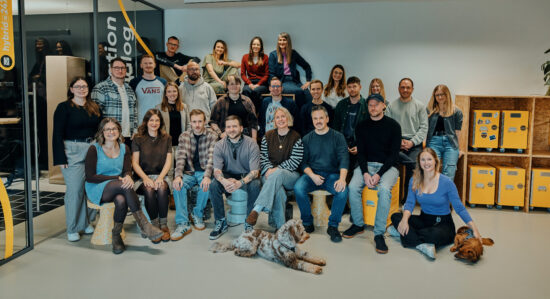 Das Team von 247GRAD sitzt und steht in mehreren Reihen im Office. Insgesamt sind 25 Personen zu sehen und zwei Bürohunde. Alle haben unterschiedliche Kleidung an und lächeln in die Kamera. Im Hintergrund sieht man einen Besprechungsraum und rollbare Flightcases in einem Holzregal.