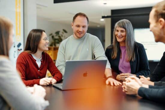 Thomas steht an einem Tisch auf der Workshopfläche im 247GRAD Office. Auf dem Tisch steht sein Macbook. Isabella und Anna stehen neben ihm und schauen sich gemeinsam die Sache an, die er ihnen zeigt.